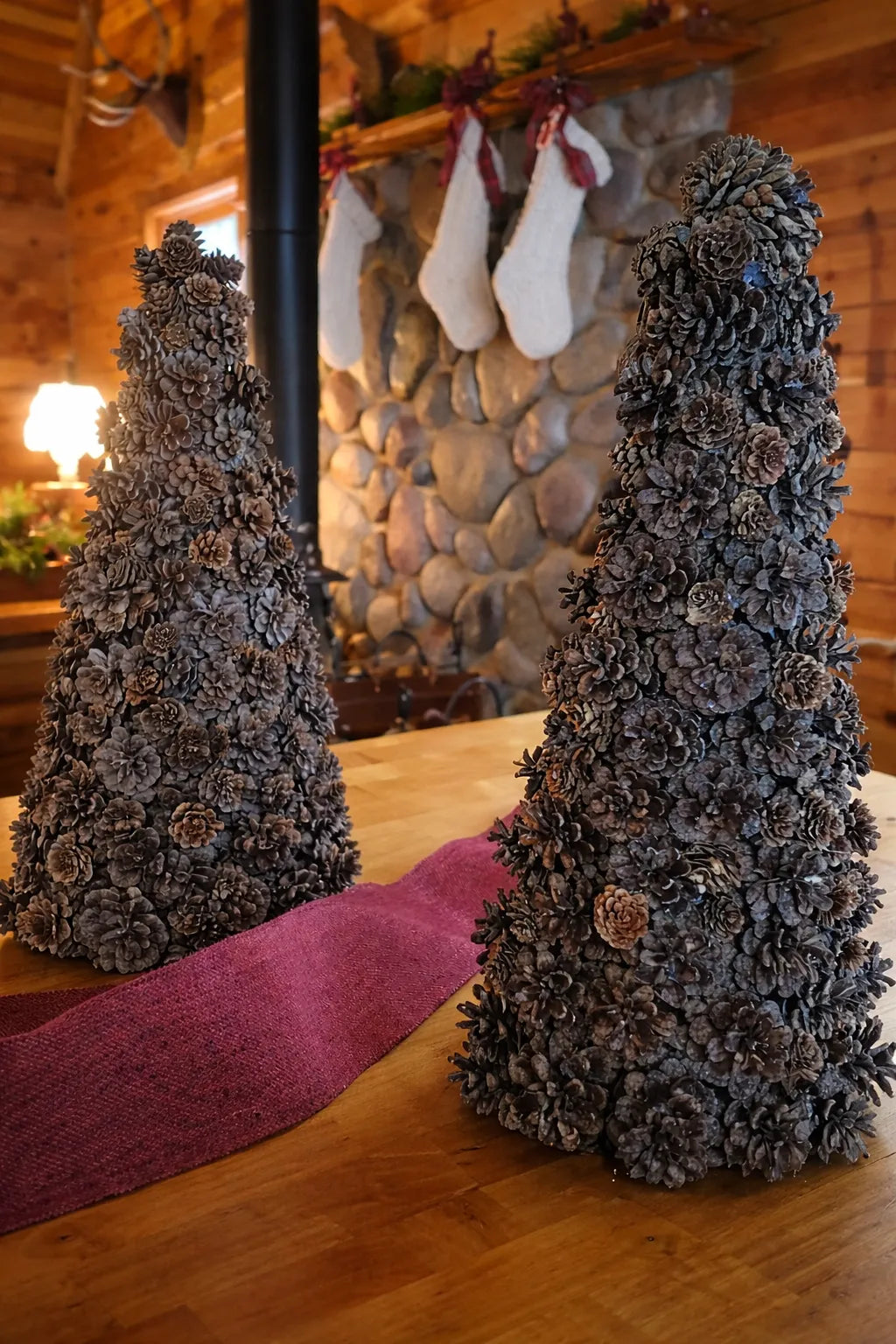 Ausable wreath company Two decorative pine cone trees on a wooden table with a stone fireplace and stockings in the background.
